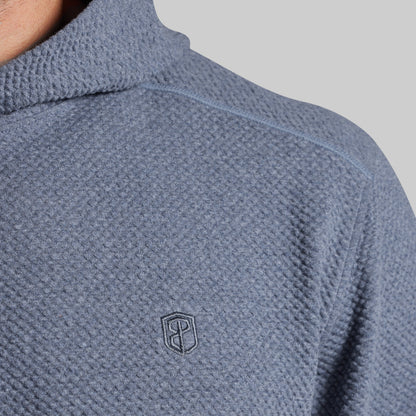 Close-up of a textured waffle-knit pullover top in a muted slate blue-gray color, featuring a small dark embroidered shield logo on the chest.