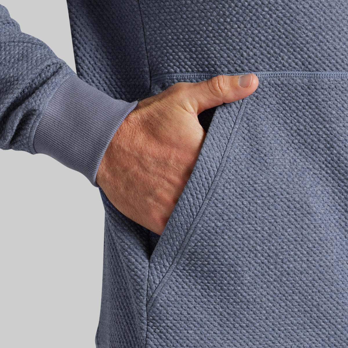 Close-up of a person wearing a textured slate blue hoodie, with a hand inserted into the side pocket.
