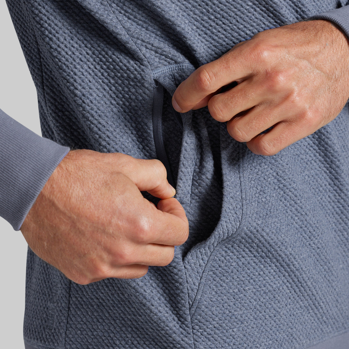Close-up of a person's hands unzipping a side pocket of a textured slate blue pullover hoodie.