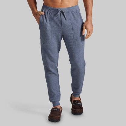 A man wearing textured slate blue jogger pants with a drawstring waist and side pockets, paired with brown boat shoes, shown from the waist down on a solid gray background.