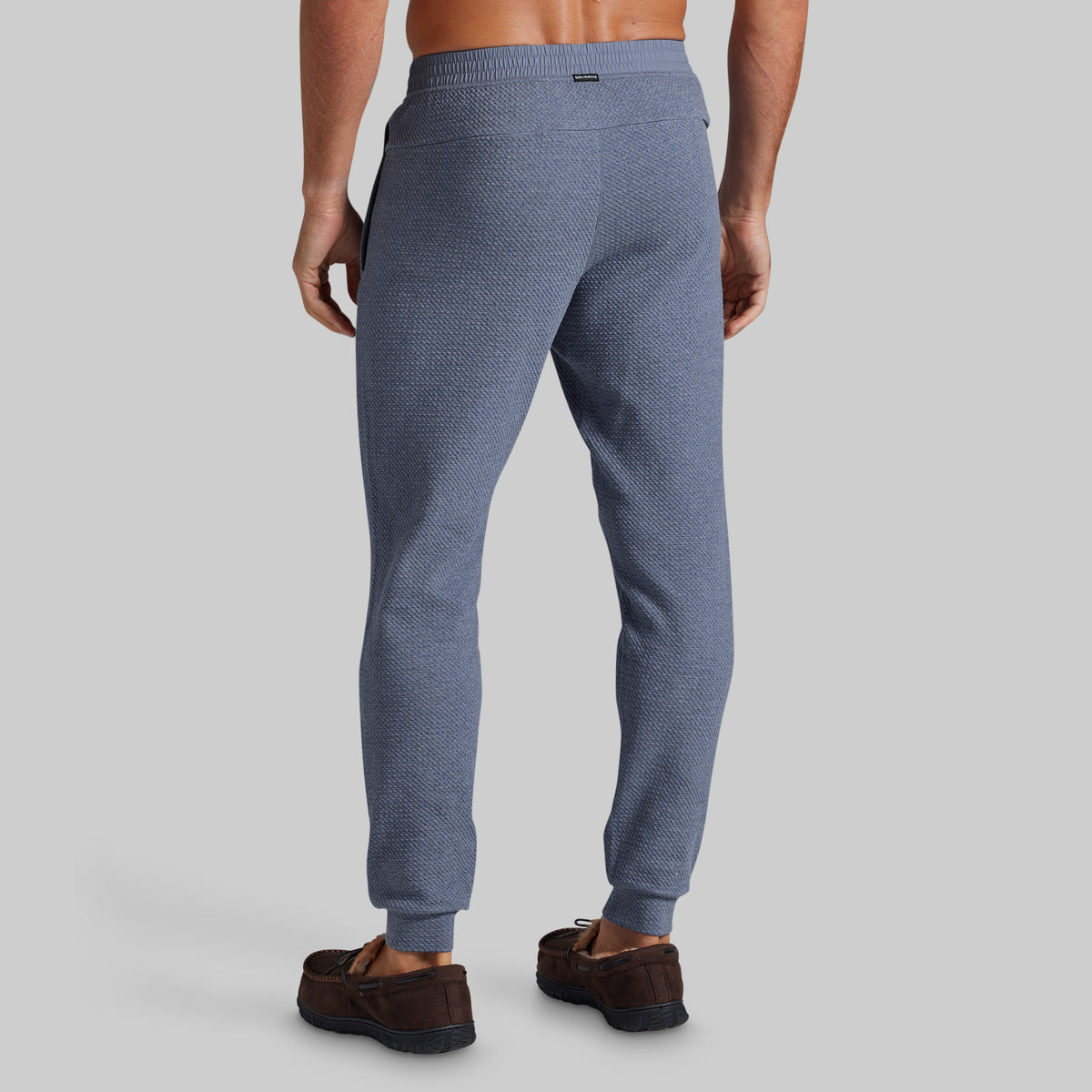 A man viewed from the back wearing textured blue jogger pants and brown suede loafers against a solid gray studio background.