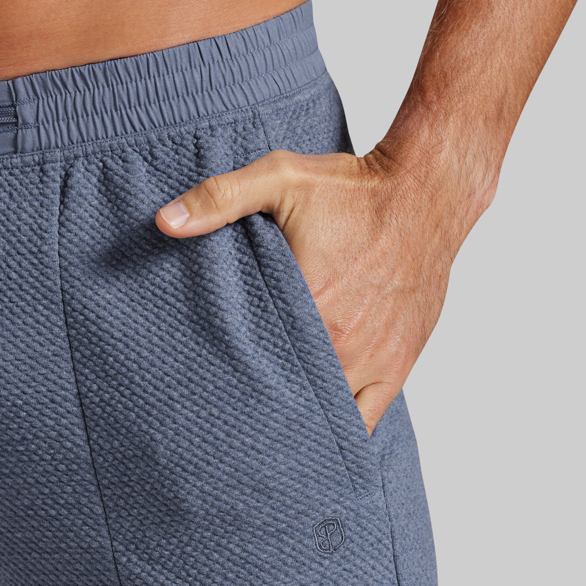 Close-up of a person wearing textured slate blue athletic joggers, showing the side pocket with a hand inserted and a small tonal logo near the lower hem.