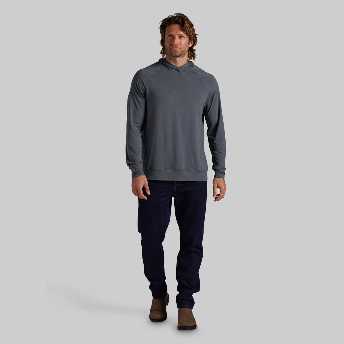 A man modeling a dark gray long-sleeve hooded sweatshirt with dark navy pants and brown boots, standing against a solid gray studio background.