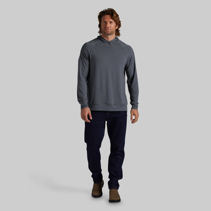 A man modeling a dark gray long-sleeve hooded sweatshirt with dark navy pants and brown boots, standing against a solid gray studio background.