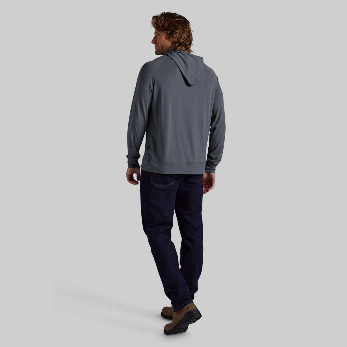 A man is shown from behind wearing a slate gray hooded sweatshirt, dark navy pants, and brown shoes, standing against a solid light gray background.
