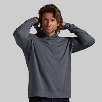 A man wearing a dark gray long-sleeve hooded shirt, posing with his hands behind his head against a solid gray background.