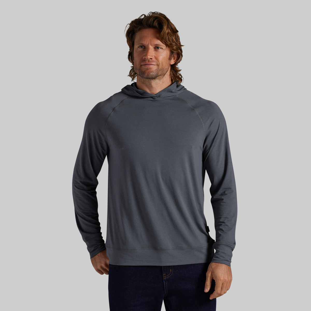 A man wearing a dark gray long-sleeve hooded sweatshirt and dark blue jeans, standing against a solid gray studio background.