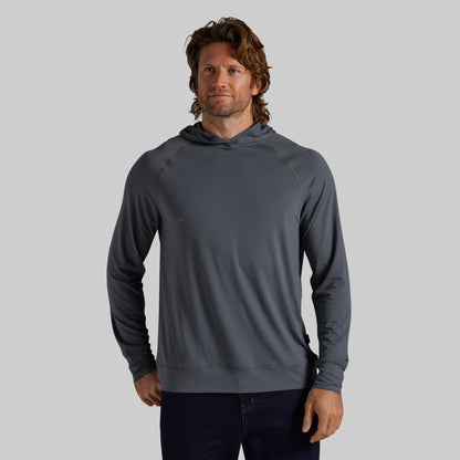 A man wearing a dark gray long-sleeve hooded sweatshirt and dark blue jeans, standing against a solid gray studio background.