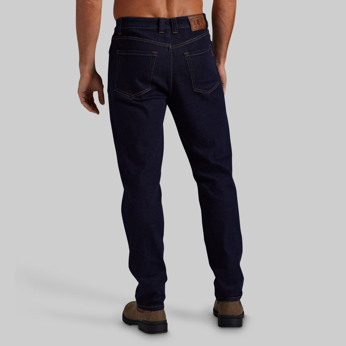 Rear view of a person wearing dark blue denim jeans and brown boots against a light gray studio background.