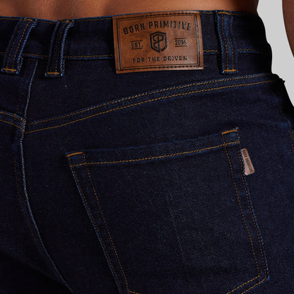 Close-up of the back waistband and pocket area of dark blue denim jeans, showing a brown leather patch that reads 'BORN PRIMITIVE EST. 2014 FOR THE DRIVEN' and a small side tag on the pocket.