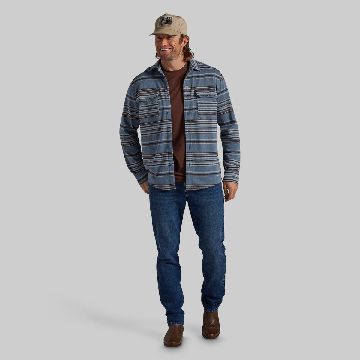 A man modeling a blue and gray striped flannel shirt over a brown t-shirt, paired with blue jeans, brown boots, and a beige baseball cap, against a solid gray background.