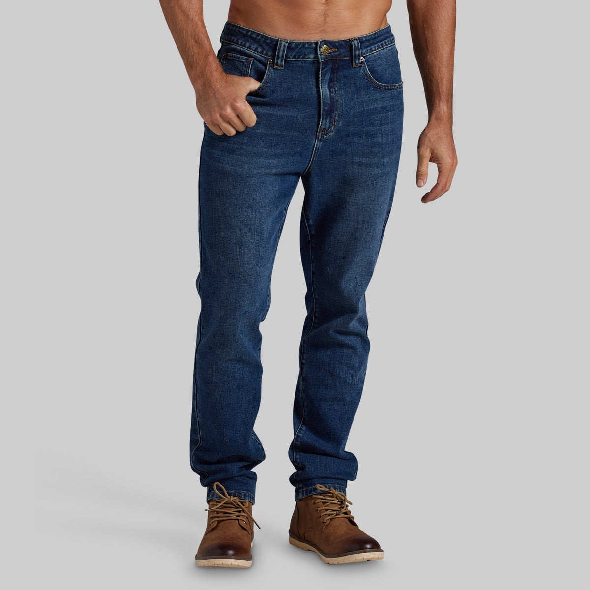 A man wearing dark blue slim-fit jeans and brown leather boots, shown from the waist down against a solid light gray background.