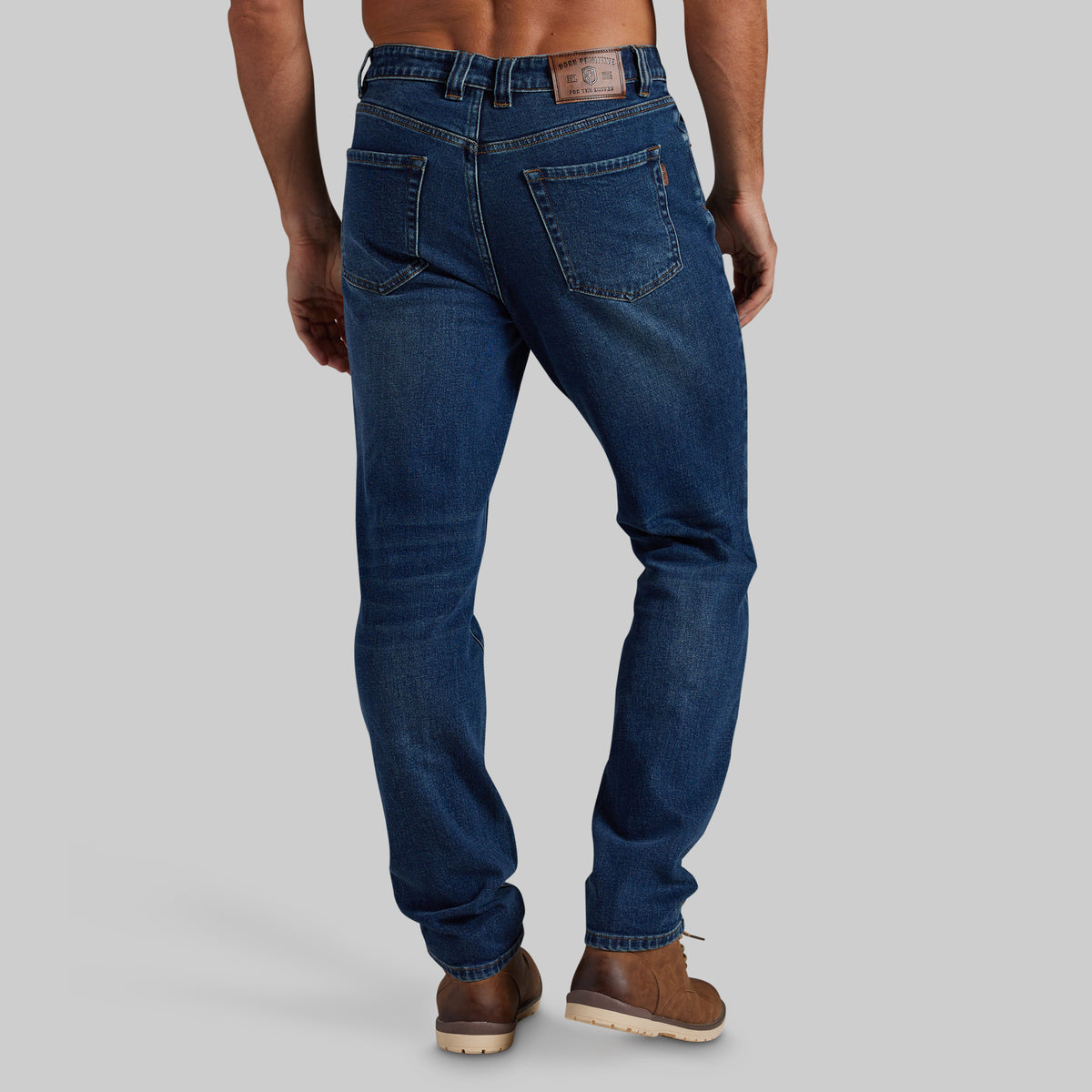 Rear view of a man wearing dark blue slim-fit jeans and brown boots against a light gray studio background.