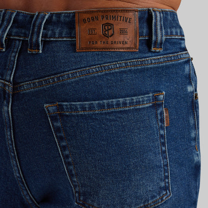 Close-up of the back waistband and pocket area of dark blue denim jeans, showing a brown leather patch that reads 'BORN PRIMITIVE EST. 2014 FOR THE DRIVEN'.