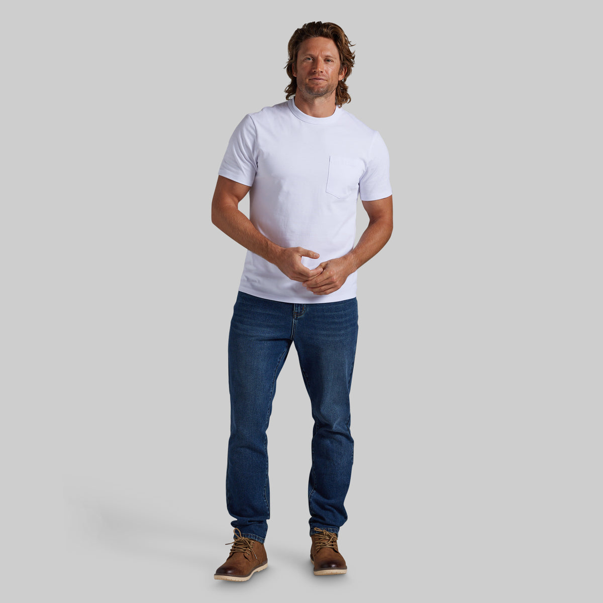 A man modeling a white short-sleeve crewneck t-shirt with a chest pocket, paired with dark blue jeans and brown boots, against a solid gray background.