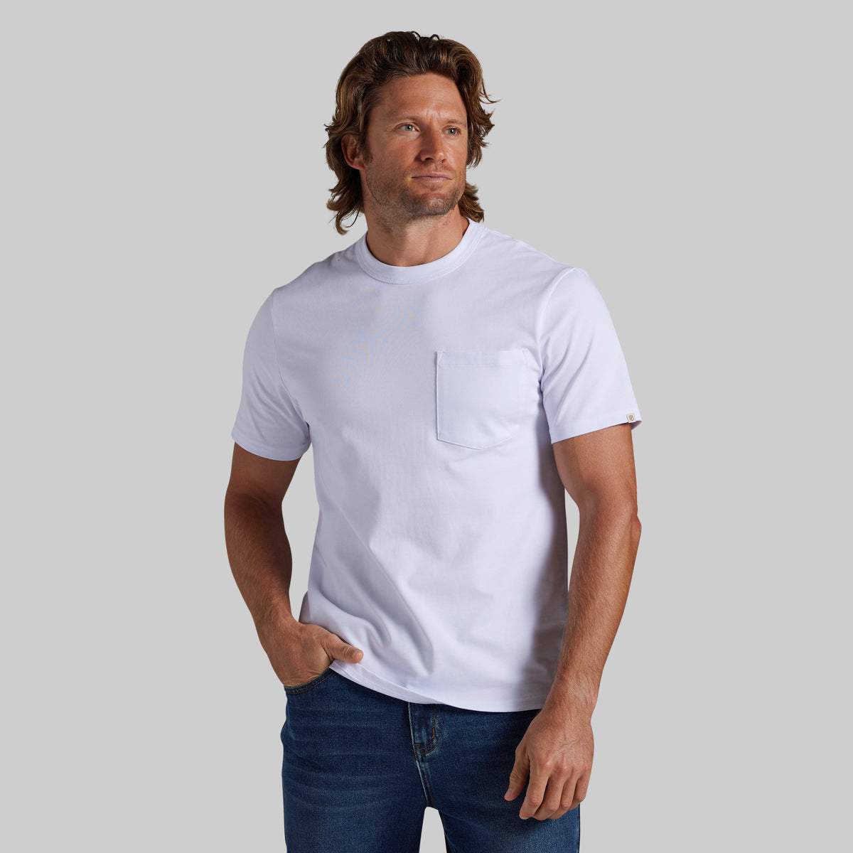 A man wearing a white short-sleeve crewneck pocket t-shirt and blue jeans, standing against a solid gray studio background.
