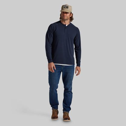 A man modeling a navy blue long-sleeve henley shirt, blue jeans, brown boots, and a tan baseball cap against a solid gray background.