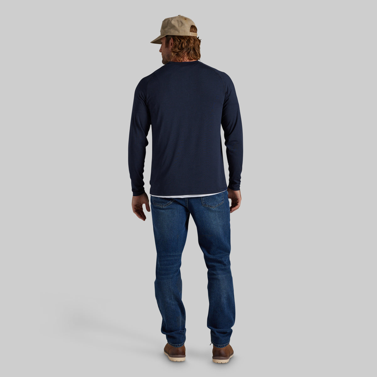 A man viewed from behind wearing a navy blue long-sleeve crewneck shirt, blue jeans, brown shoes, and a beige baseball cap, set against a solid light gray background.