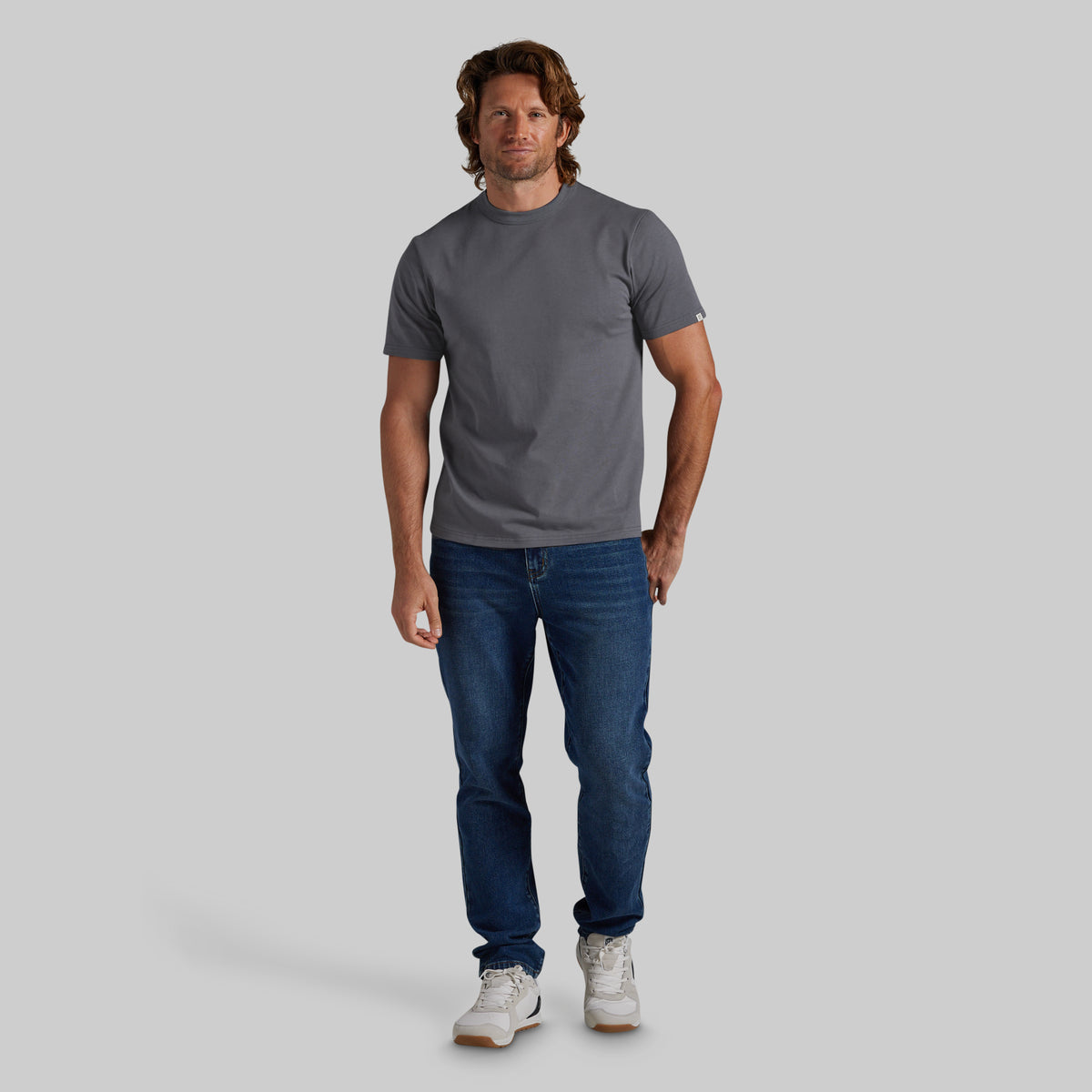 A man modeling a dark gray crewneck t-shirt, blue jeans, and white sneakers against a solid light gray background.