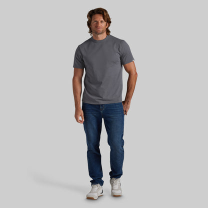A man modeling a dark gray crewneck t-shirt, blue jeans, and white sneakers against a solid light gray background.