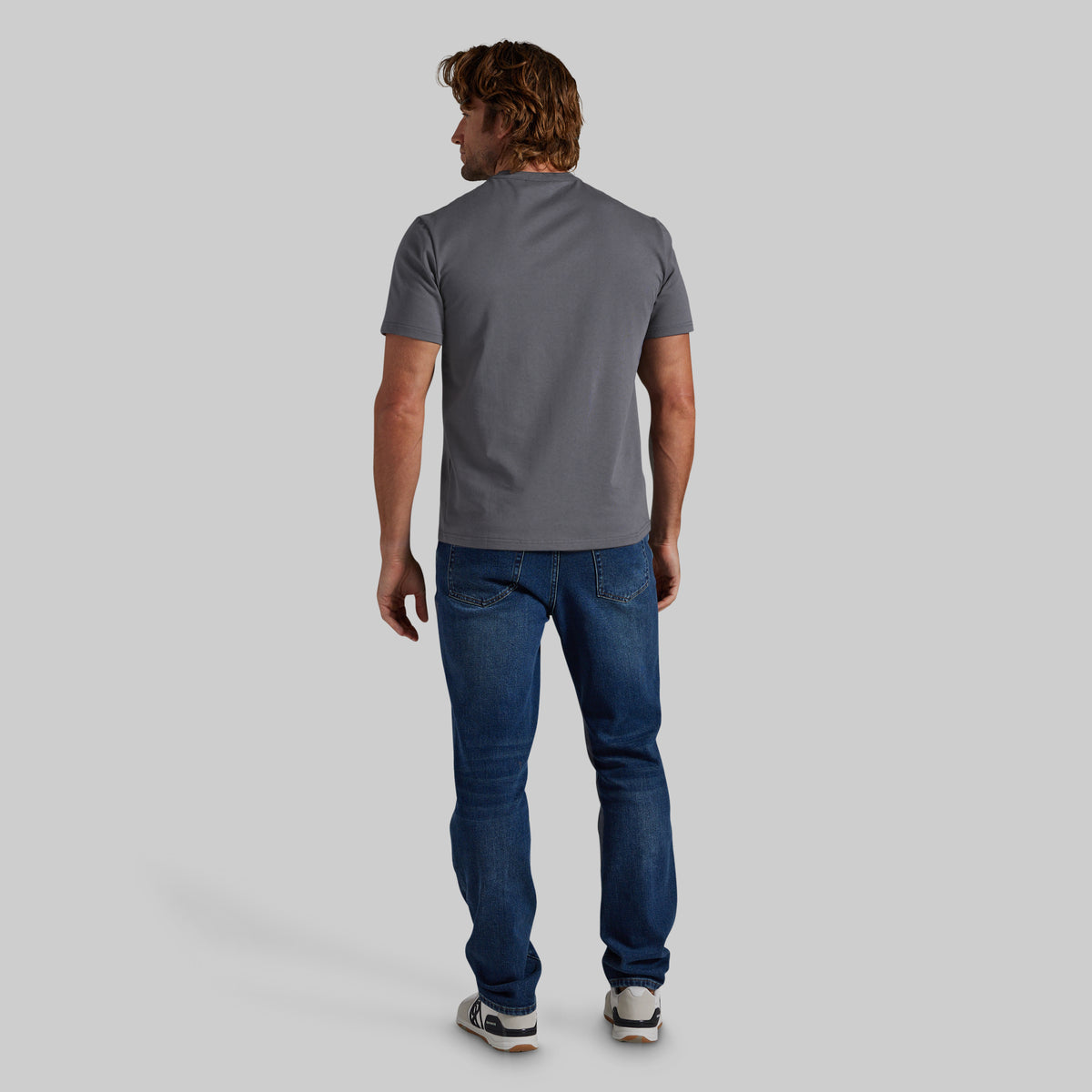 A man viewed from behind wearing a dark gray short-sleeve t-shirt, blue jeans, and white sneakers, standing against a solid gray studio background.