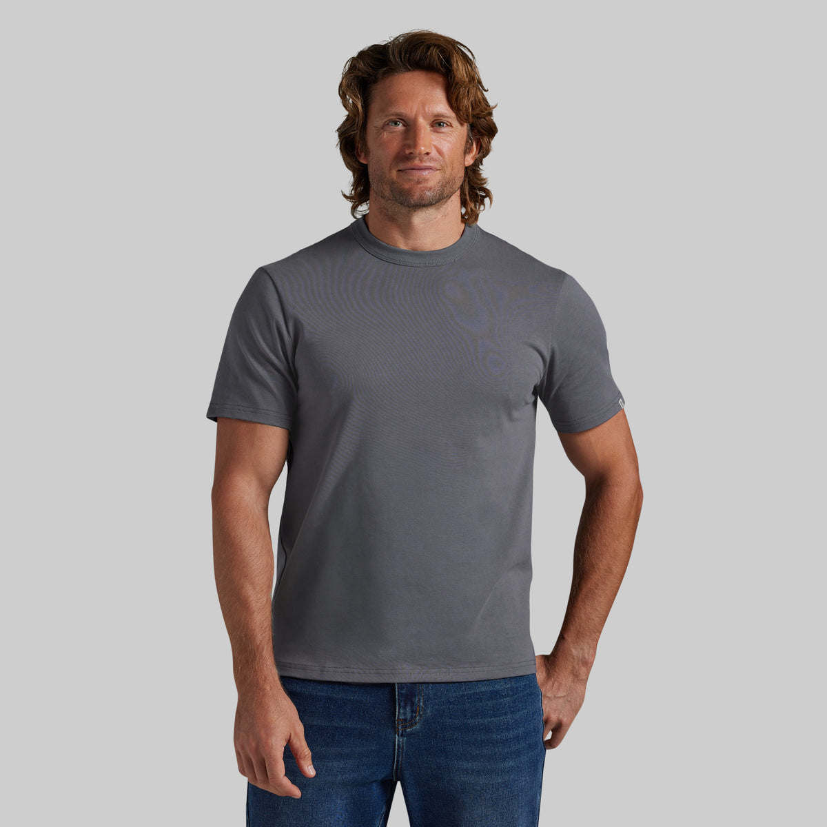 A man wearing a dark gray crewneck t-shirt paired with blue jeans, standing against a solid gray background.