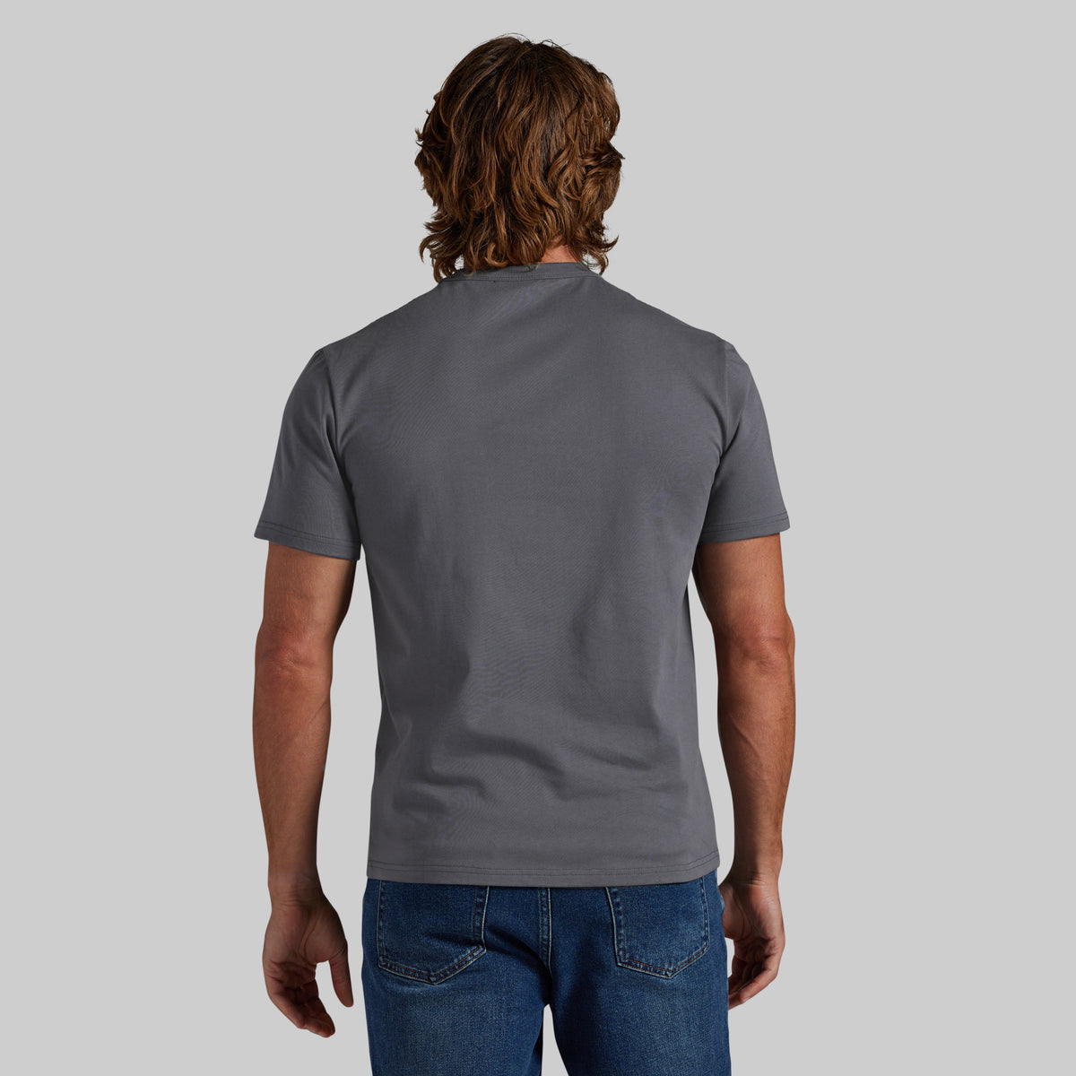 A man viewed from behind wearing a dark gray crewneck t-shirt and blue jeans, standing against a solid light gray background.