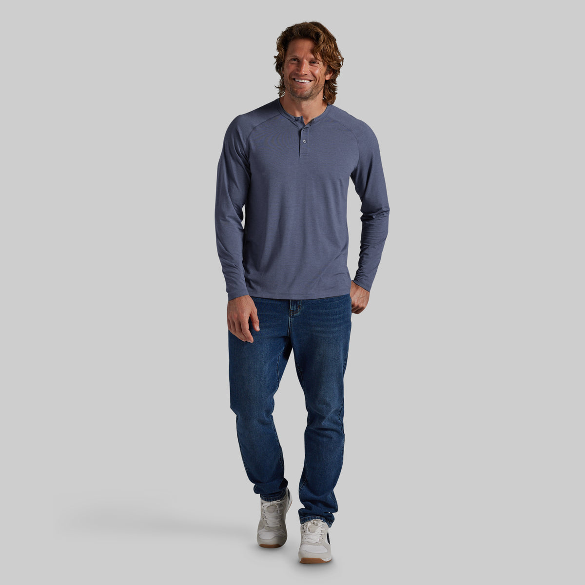 A man wearing a slate blue long-sleeve henley shirt, dark blue jeans, and white sneakers, standing against a solid gray background.