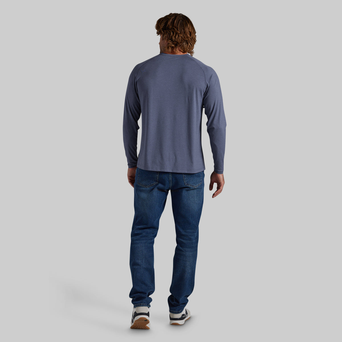 A man viewed from behind wearing a slate blue long-sleeve shirt and blue jeans, standing against a solid gray background.