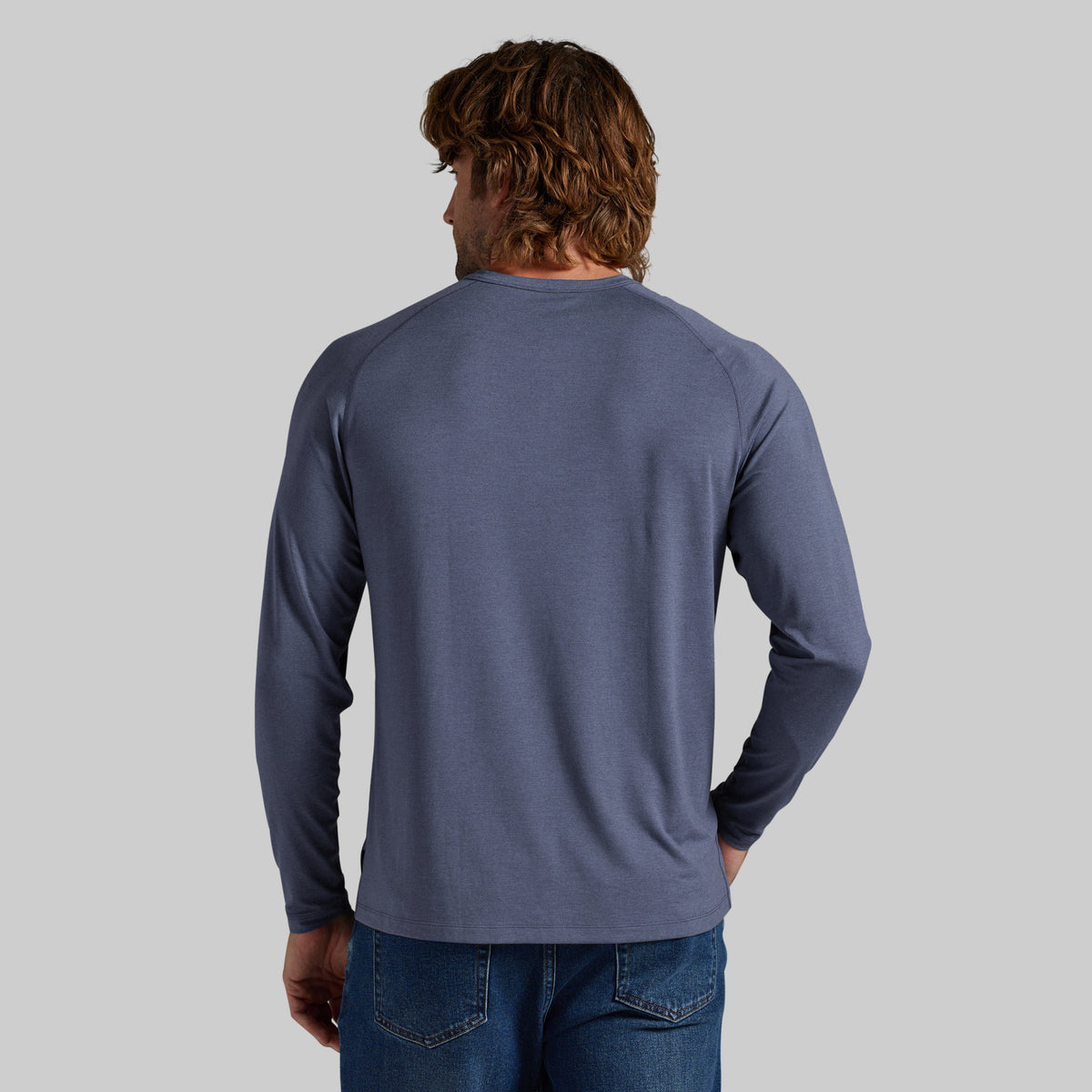 A man viewed from behind wearing a slate blue long-sleeve raglan t-shirt and blue jeans, set against a solid light gray background.