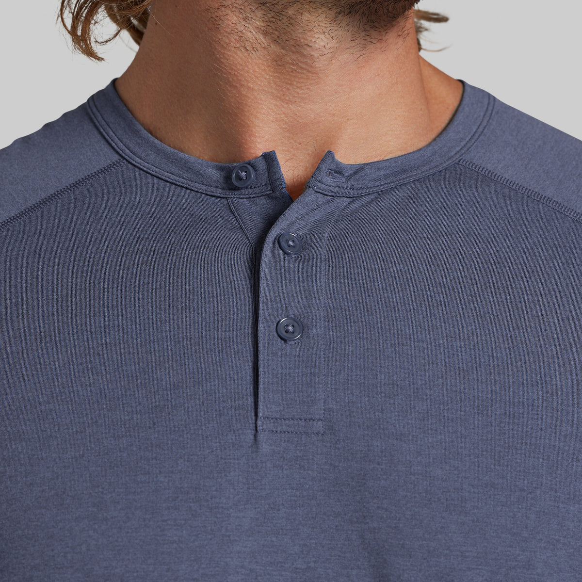 Close-up of a man wearing a heathered slate blue henley shirt with a three-button placket, showing the neck, collar, and chest area against a light gray background.