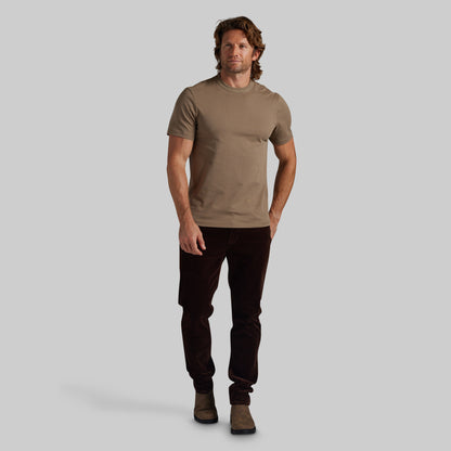 A man modeling a short-sleeve crewneck t-shirt in a muted taupe-brown color, paired with dark brown corduroy pants and brown boots, against a solid light gray background.