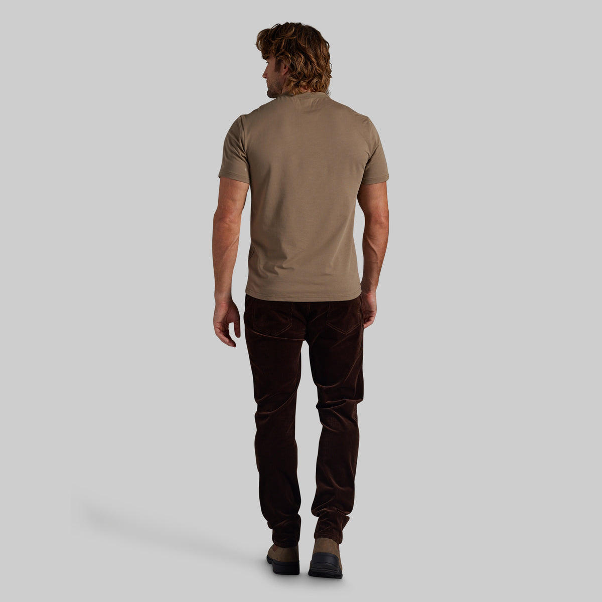 A man shown from behind wearing a short-sleeve taupe t-shirt and dark brown corduroy trousers, standing against a solid light gray background.