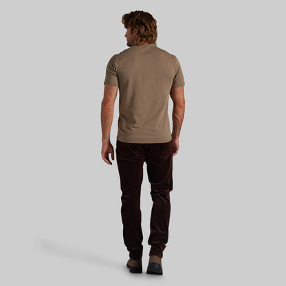 A man shown from behind wearing a short-sleeve taupe t-shirt and dark brown corduroy trousers, standing against a solid light gray background.