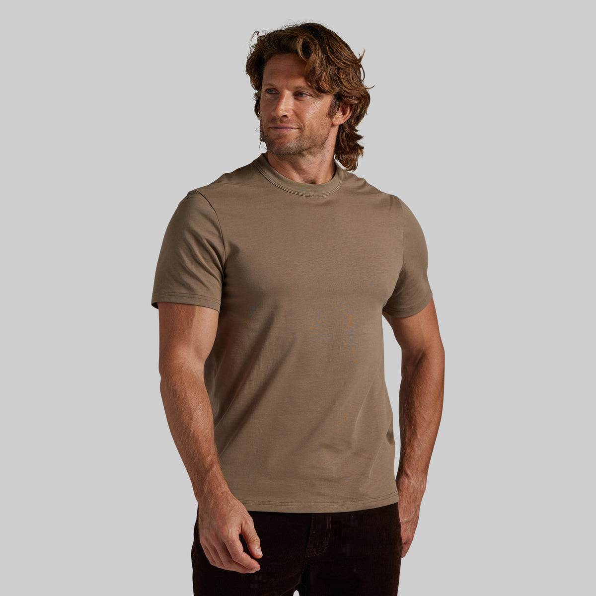 A man wearing a short-sleeve crewneck t-shirt in a muted taupe-brown color, paired with dark pants, standing against a solid light gray background.