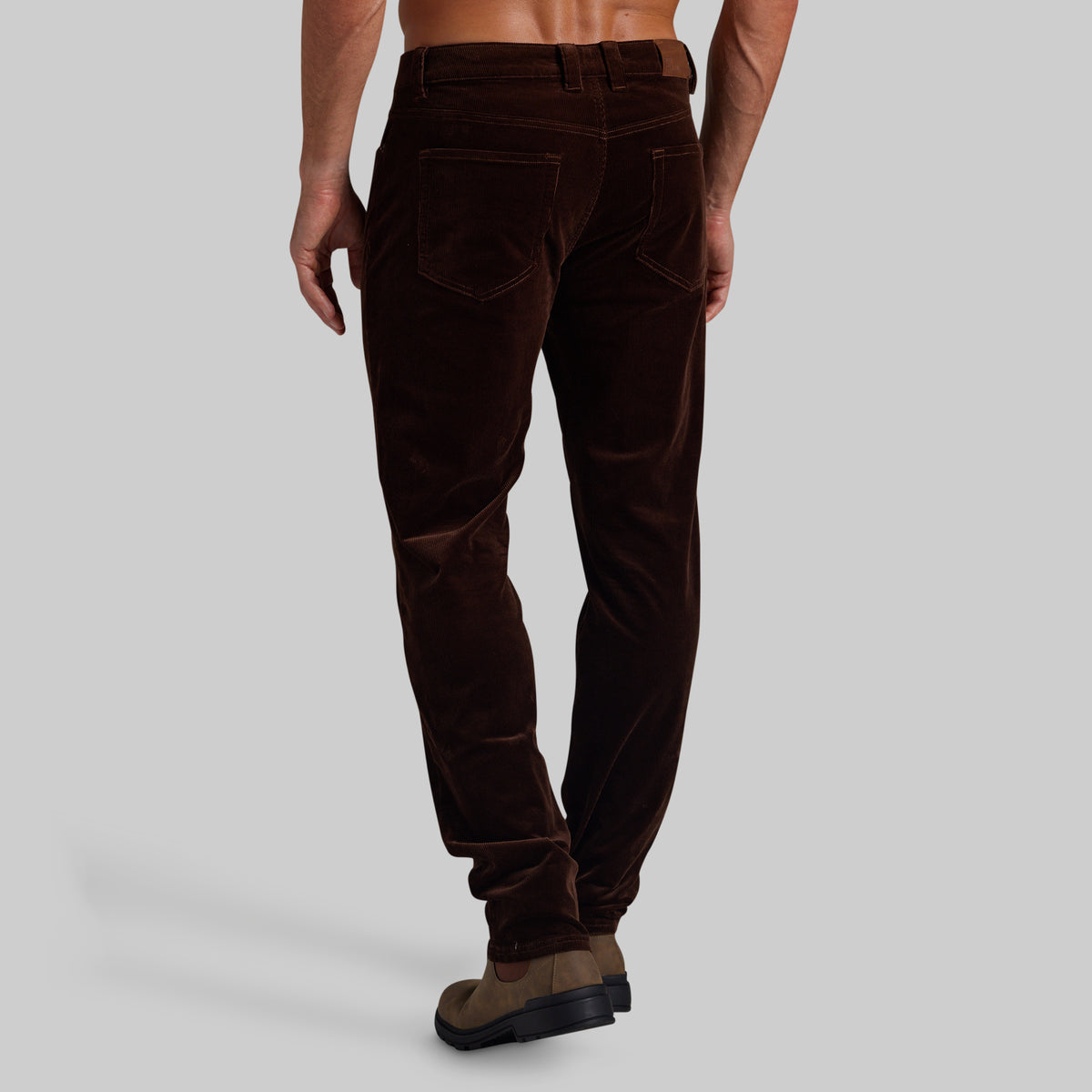 A man wearing dark brown corduroy trousers, shown from a rear three-quarter angle against a solid light gray background.