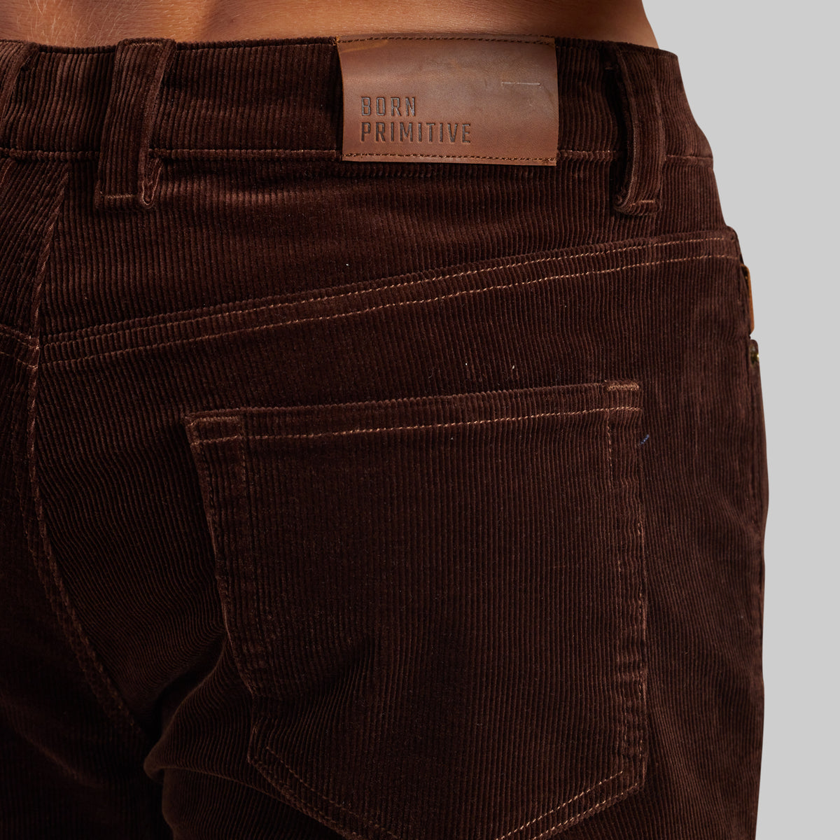 Close-up of the back waistband and pocket of dark brown corduroy pants, showing a brown leather patch with the brand name 'BORN PRIMITIVE'.