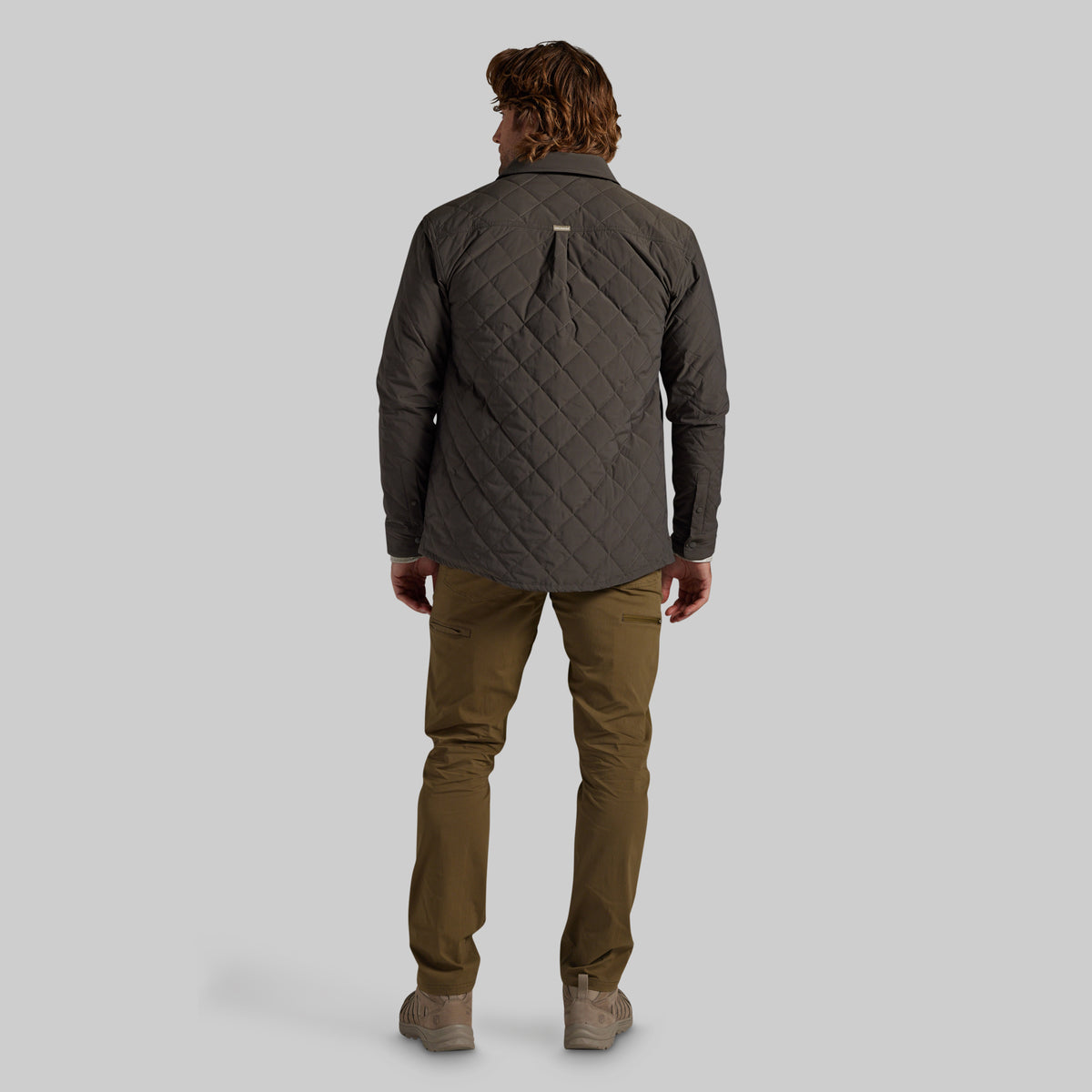 A man viewed from behind wearing a dark gray quilted jacket, olive green pants, and beige shoes, standing against a solid gray studio background.