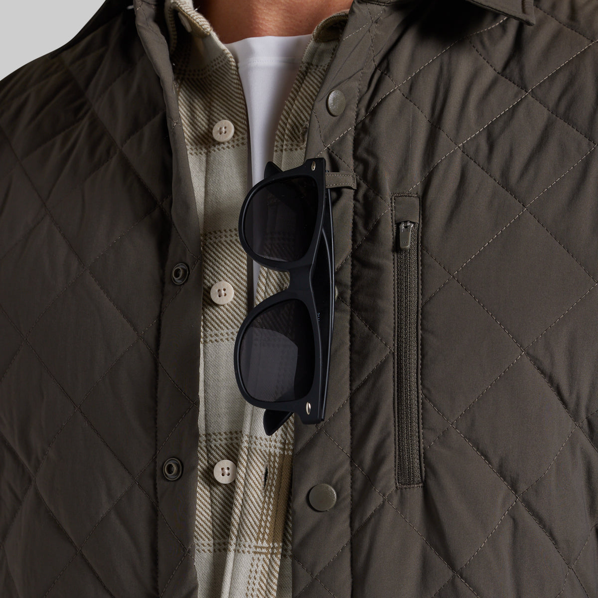 Close-up of a person wearing a dark olive quilted jacket over a beige plaid button-up shirt and white undershirt, with black sunglasses hanging from the chest pocket.