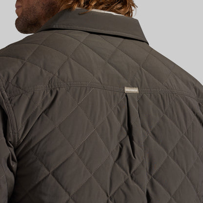 Close-up back view of a man wearing a dark olive quilted jacket, showing the diamond stitching and a small rectangular brand tag on the upper back against a light gray background.