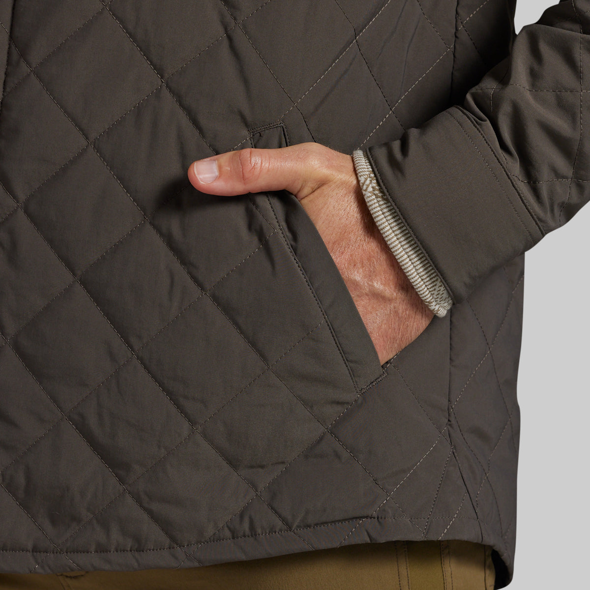 Close-up of a dark brown diamond-quilted jacket with a side pocket, showing a hand in the pocket and the ribbed inner cuff.