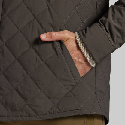 Close-up of a dark brown diamond-quilted jacket with a side pocket, showing a hand in the pocket and the ribbed inner cuff.
