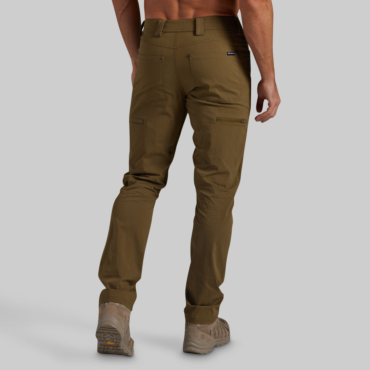 A man shown from the back wearing olive brown cargo pants and tan hiking boots against a light gray studio background.