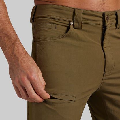 Close-up of a person wearing olive green chino pants, showing the side pocket and hand pulling the pocket flap open, against a light gray studio background.