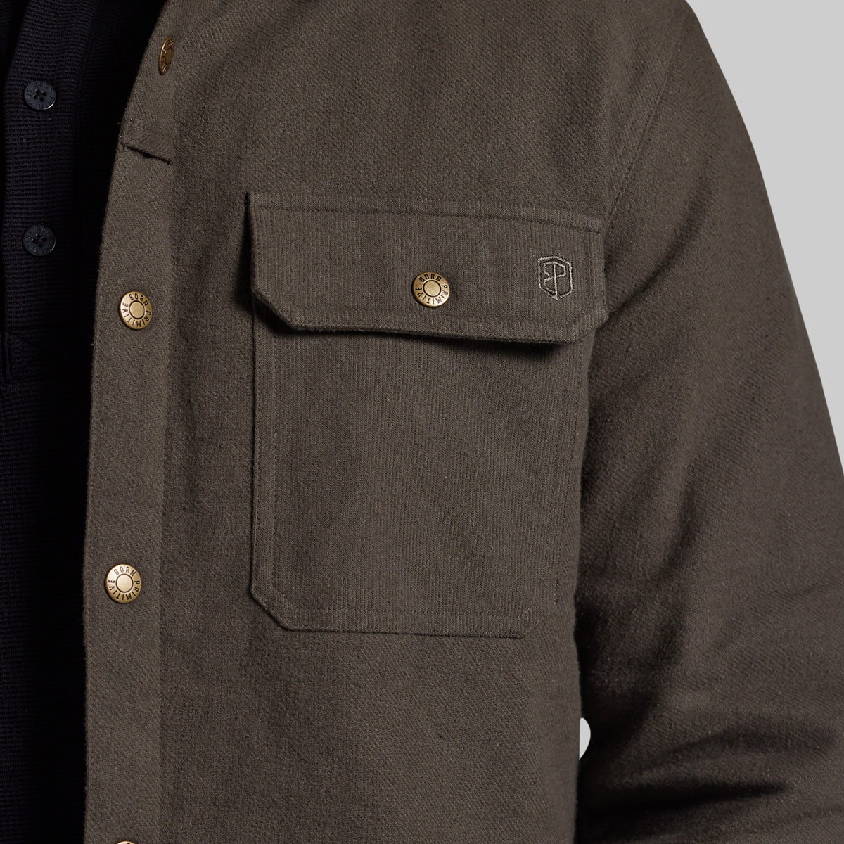 Close-up of a dark olive brown textured overshirt jacket with brass buttons and a small embroidered shield logo on the chest pocket, worn over a dark shirt.