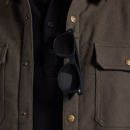 Close-up of a person wearing an olive brown textured jacket over a black knit shirt, with black sunglasses resting on the chest pocket.