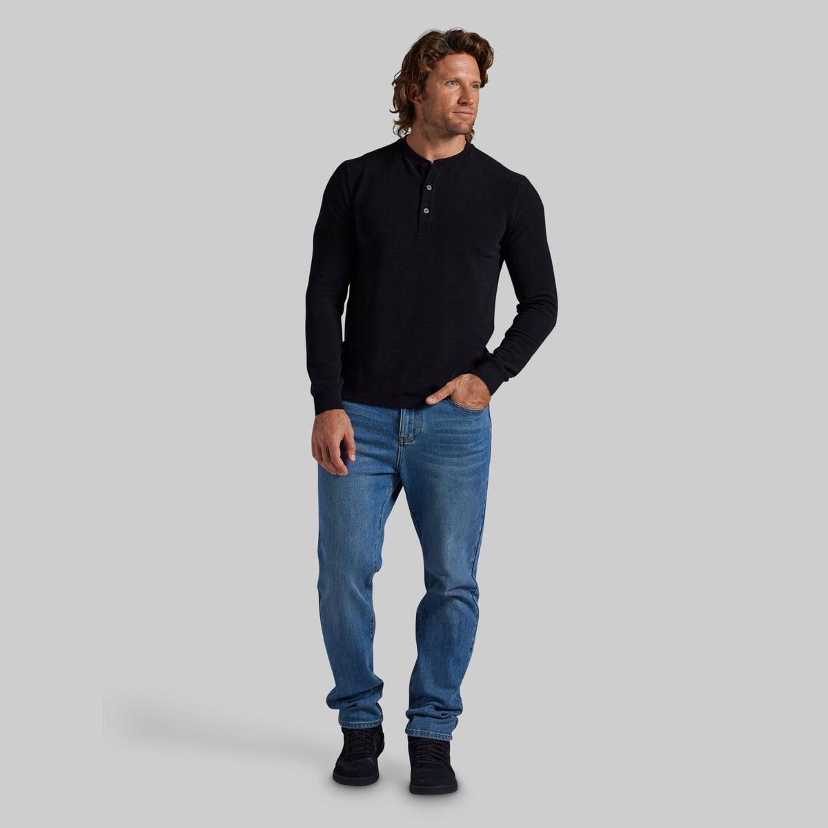 A man modeling a black long-sleeve henley shirt with blue jeans and black shoes, standing against a solid gray studio background.