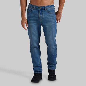 A person modeling a pair of medium blue slim-fit jeans with black sneakers against a solid light gray studio background.