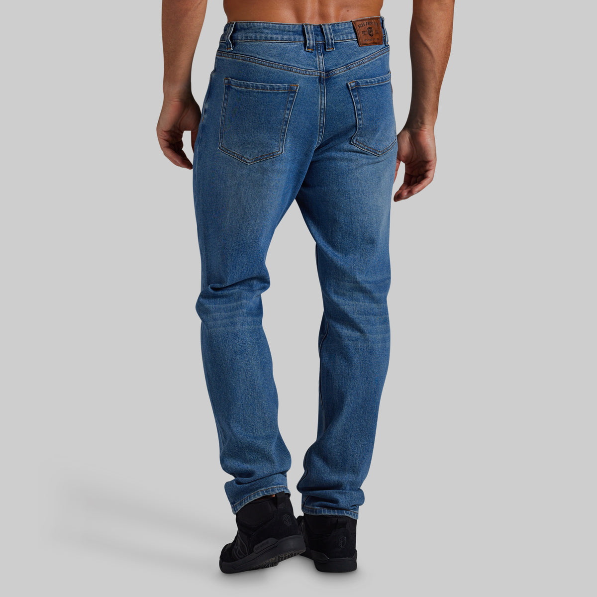 Rear view of a man wearing blue slim-fit jeans with black boots, shown against a solid gray studio background.