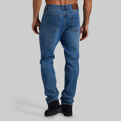 Rear view of a man wearing blue slim-fit jeans with black boots, shown against a solid gray studio background.